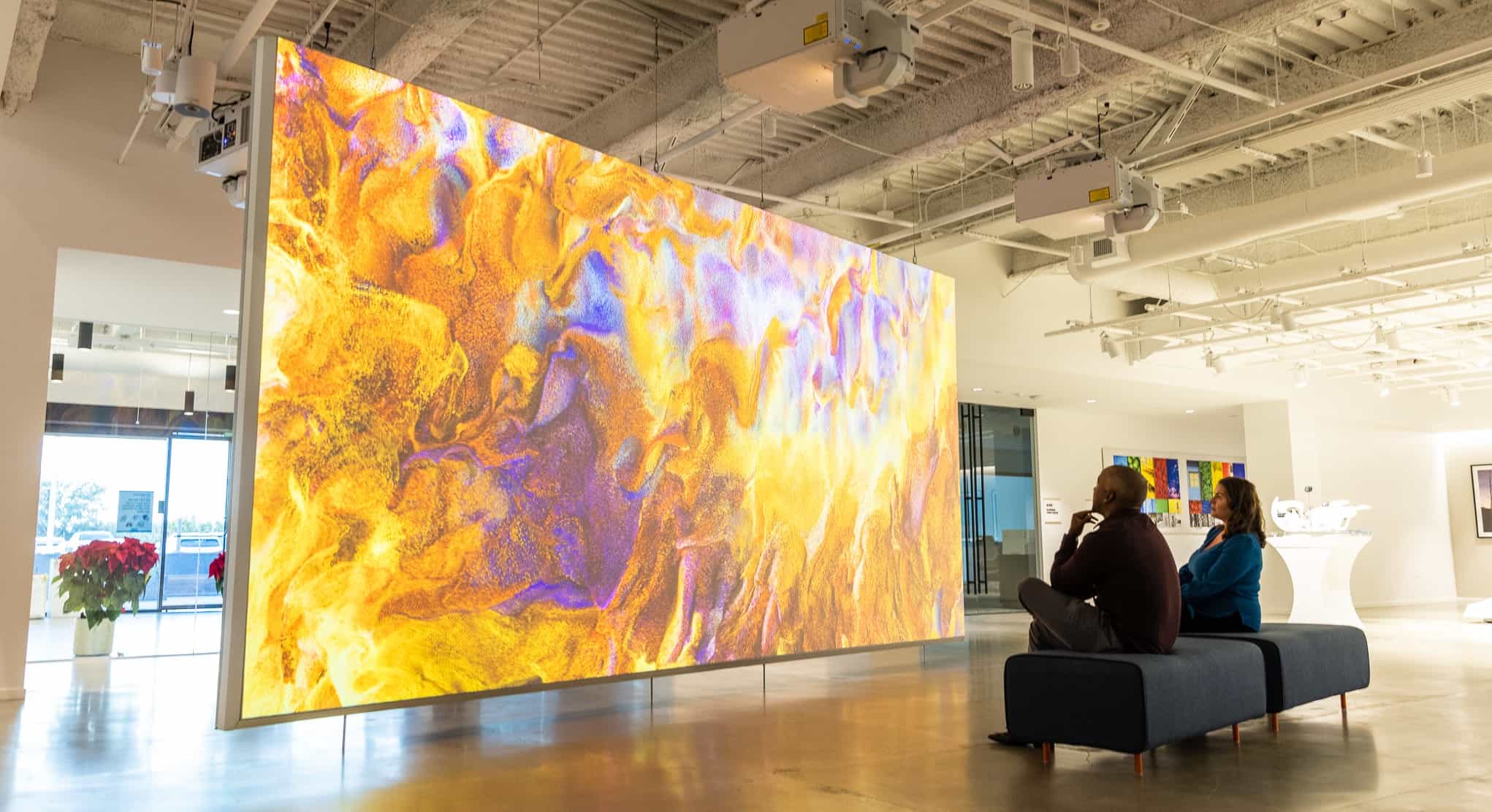 Two people on blue benches in a gallery watch a vibrant digital display with abstract patterns; industrial ceiling above.