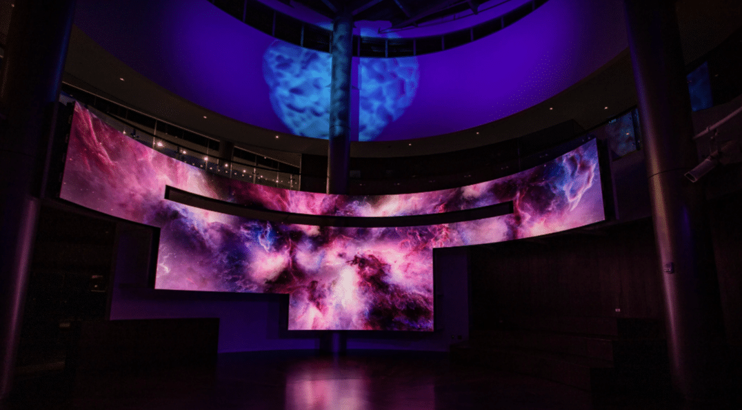 A large digital screen shows a vibrant nebula in purples, pinks, and blues. Purple ambient lighting enhances the immersive vibe.