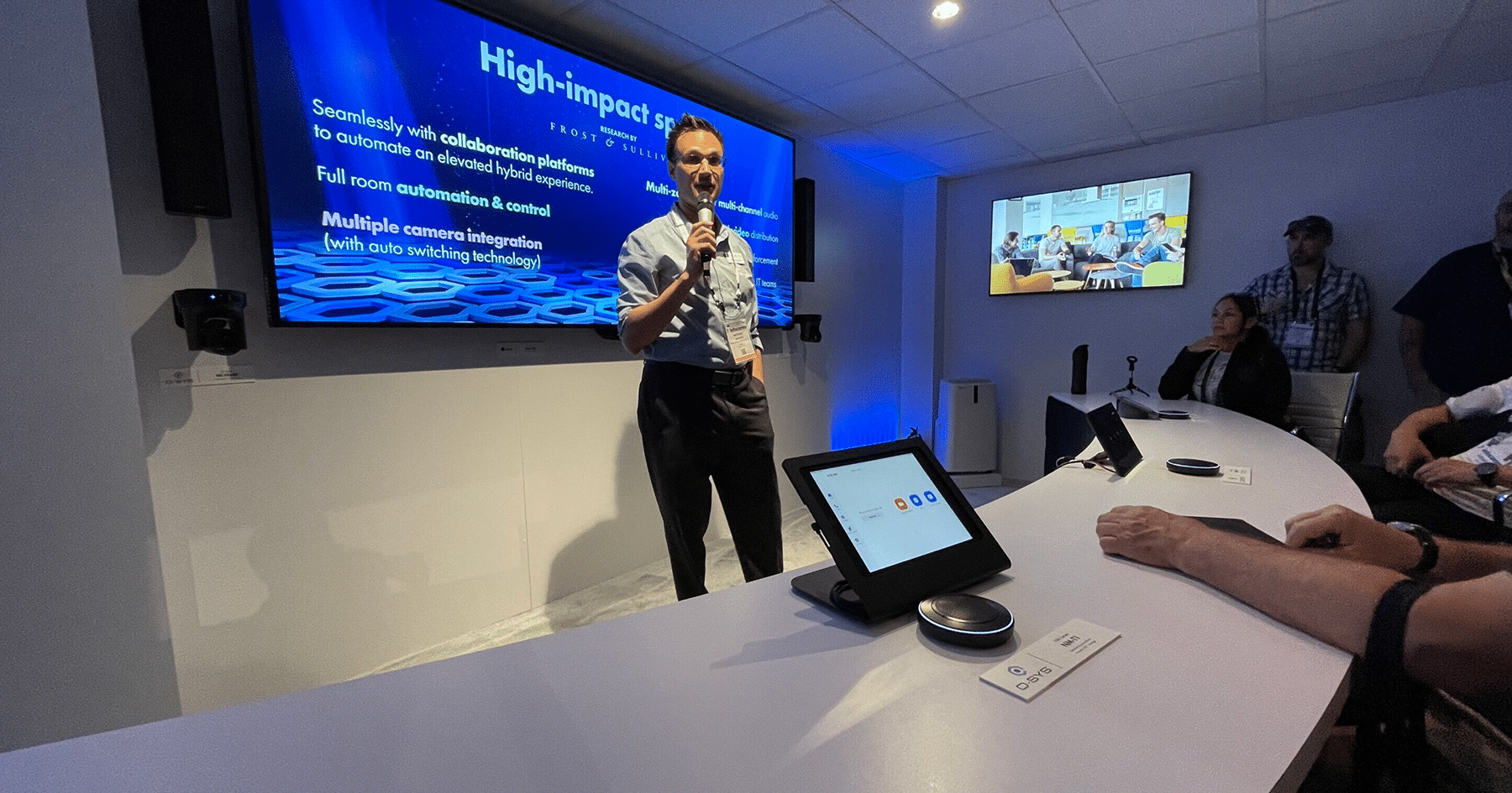 A man speaks at a curved desk with screens; attendees watch a high-impact solutions presentation, tablets on desk.