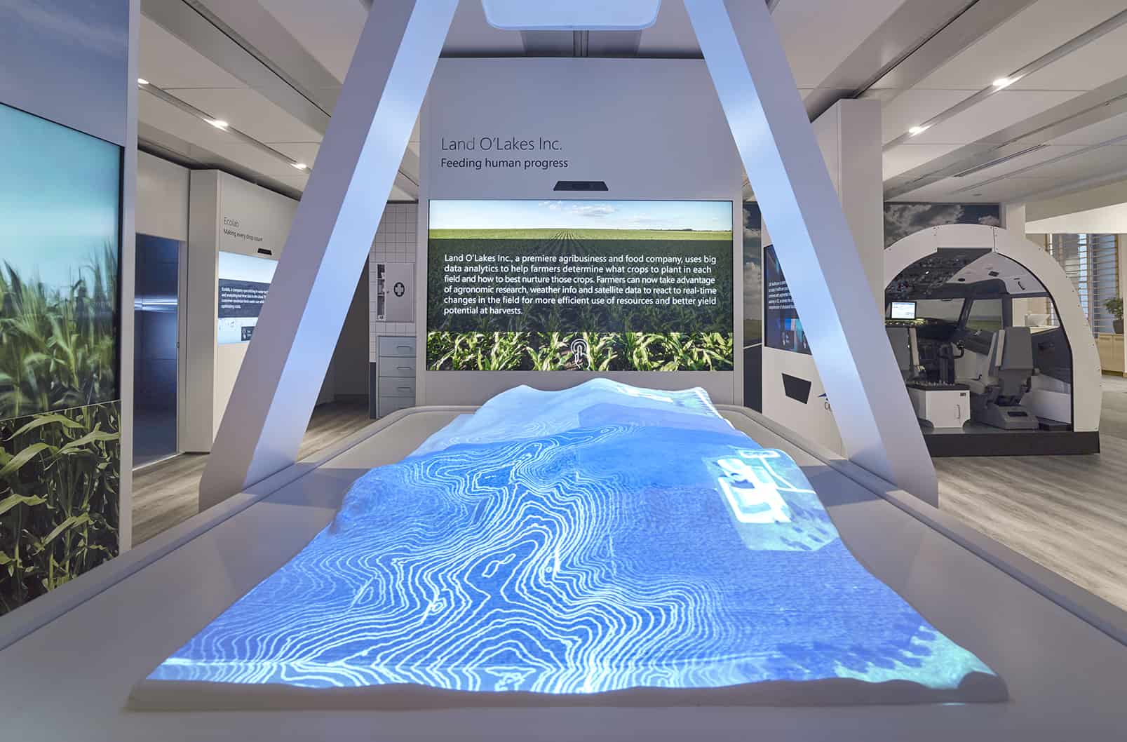 Futuristic room with a contoured table, blue-white topographic display, cornfield images, and info panels under ambient lighting.