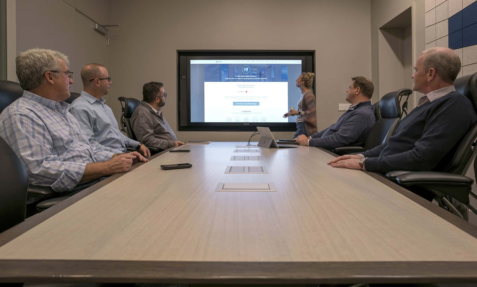 Five people sit at a conference table, screen display; one stands pointing, modern well-lit room.