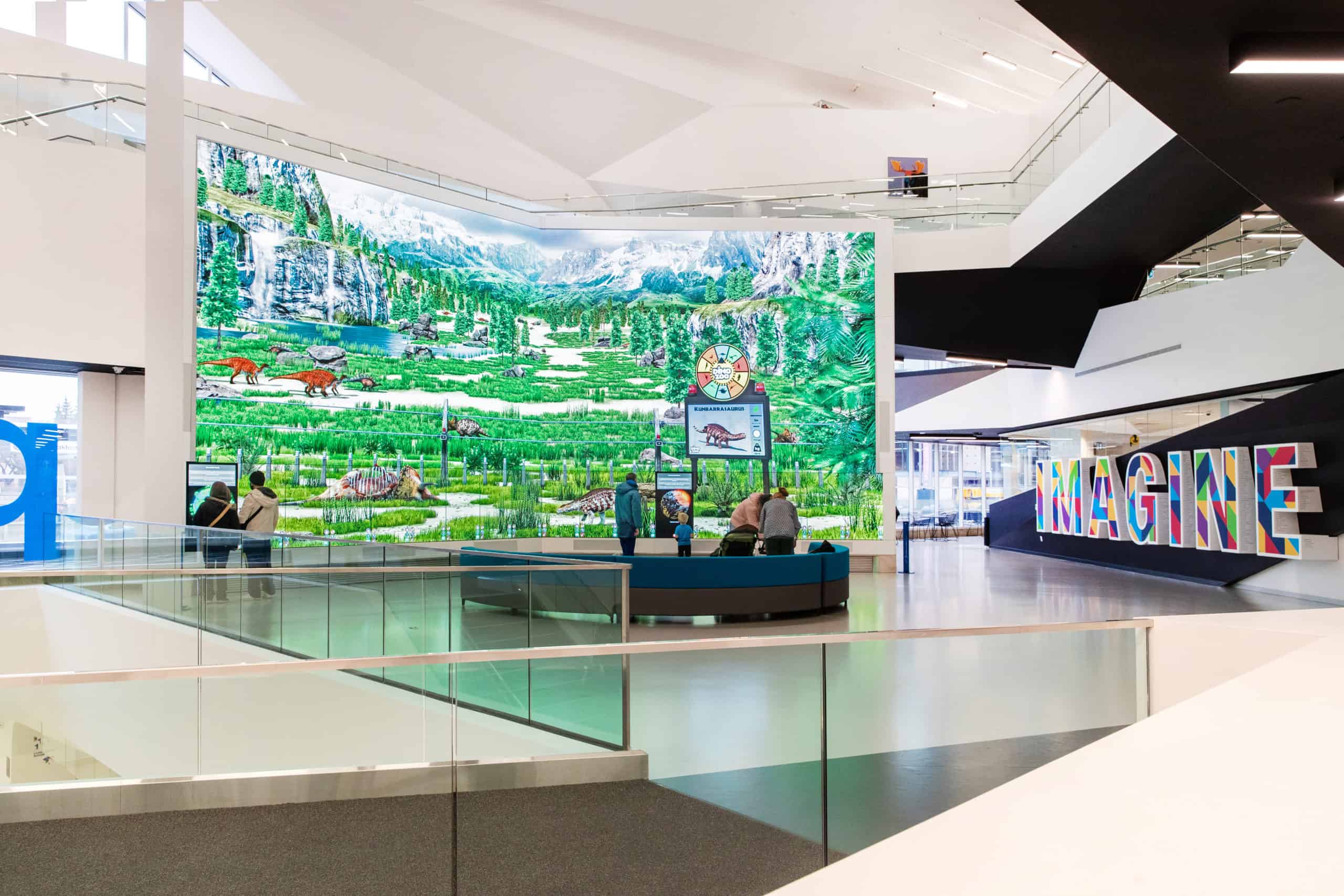 A spacious interior features a vibrant mural of nature. People observe. IMAGINE spells out in colorful letters. Bright, modern room.