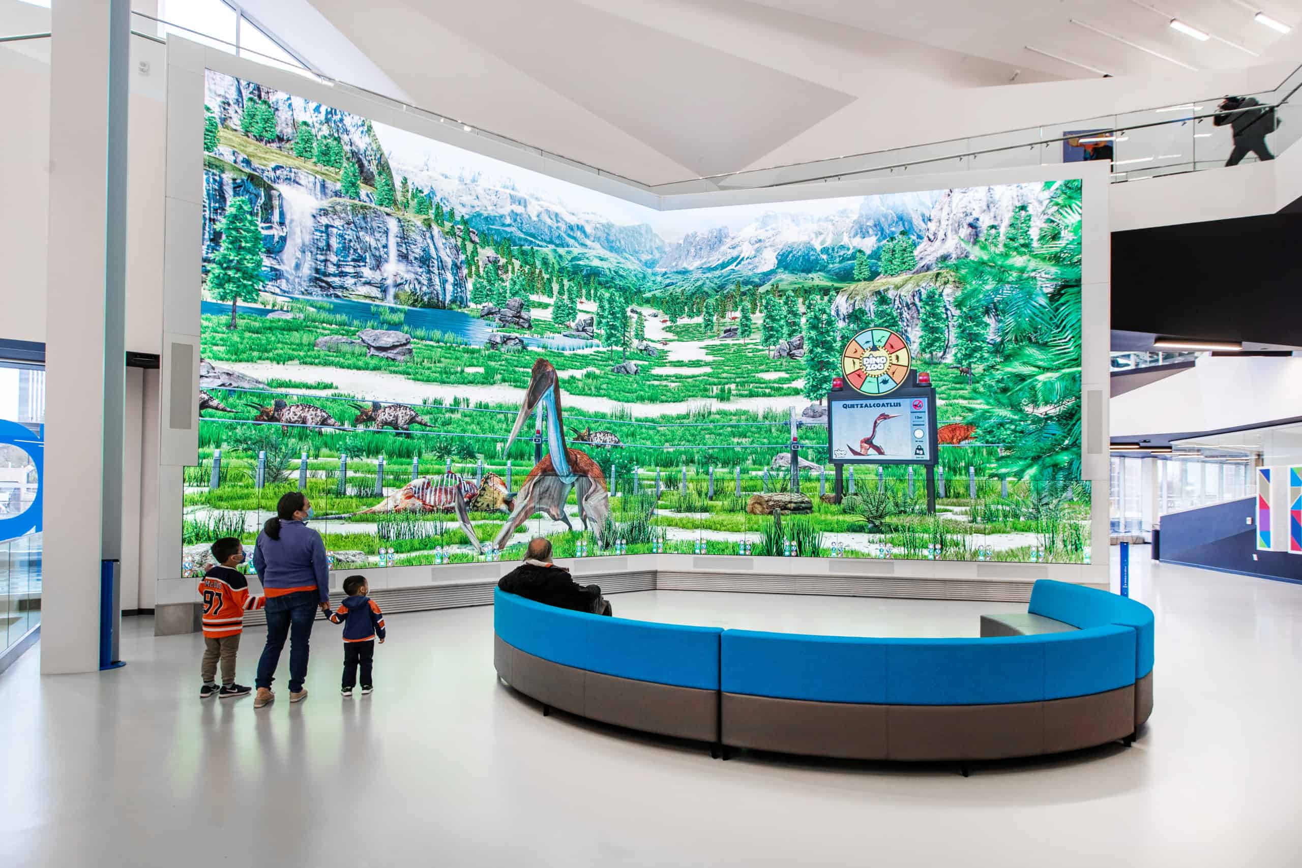 A group stands by a curved bench in a gallery, viewing a large display of dinosaurs, mountains, and greenery.