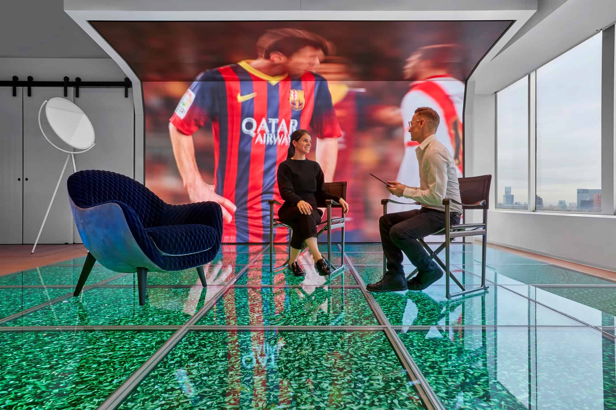 Two people sit in a modern room with a screen showing a Barcelona player; green floor, blue chair by window with city view.