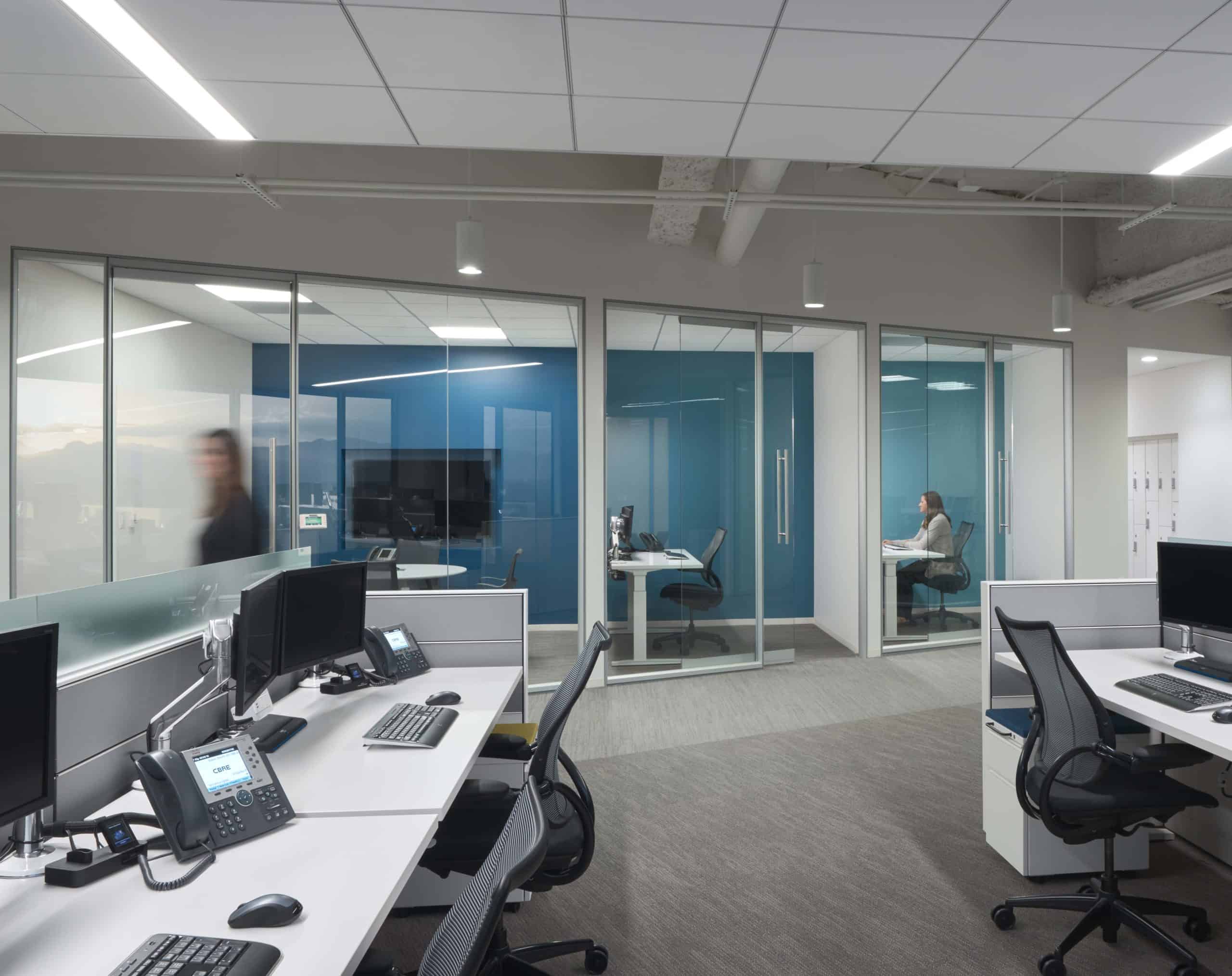 Modern open office with white desks, glass meeting room, blue accent walls; one person seated inside, another walking outside.