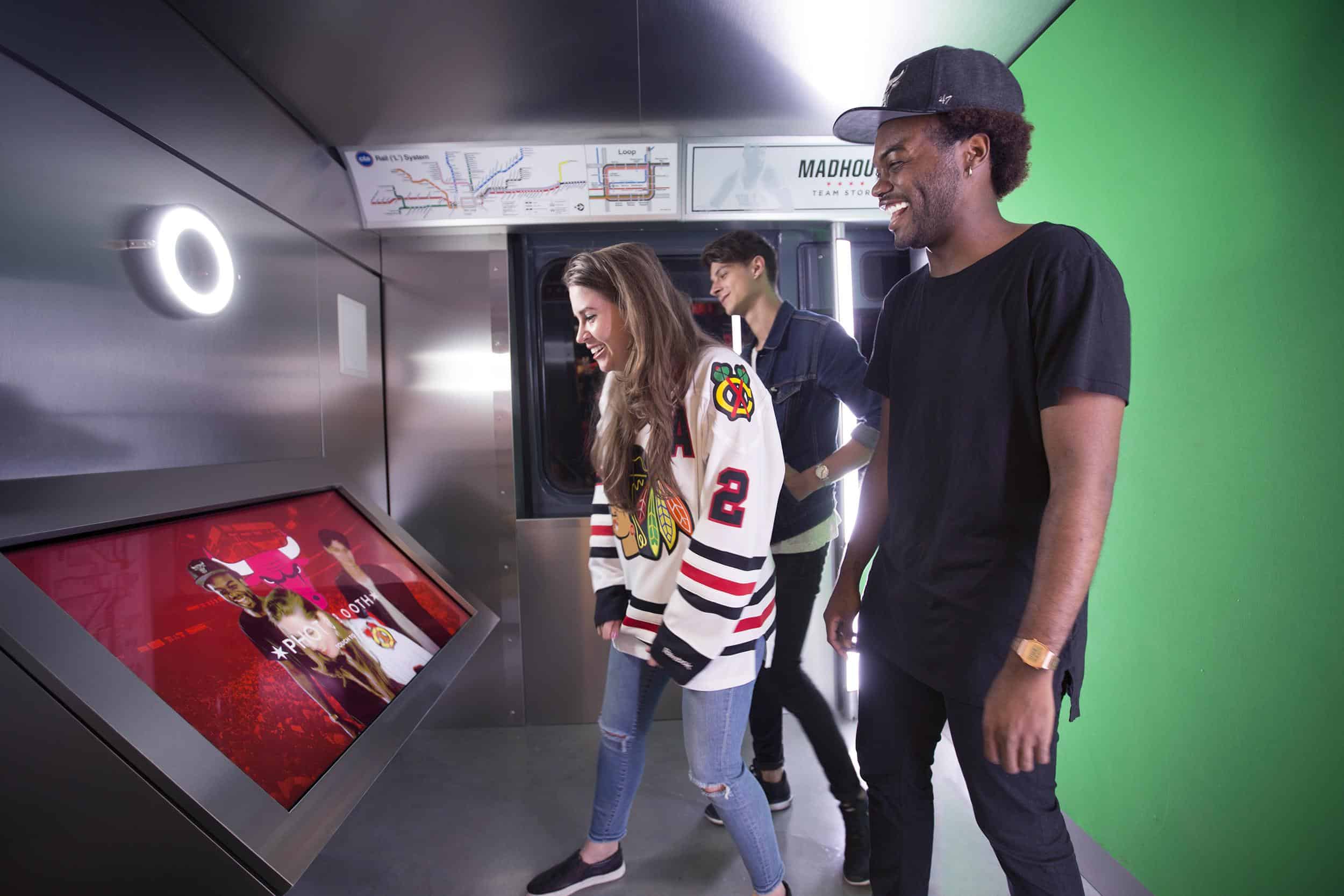 Three people smile, interacting with a touchscreen. One in a sports jersey. Gray floor, green screen backdrop, ring light, map overhead.