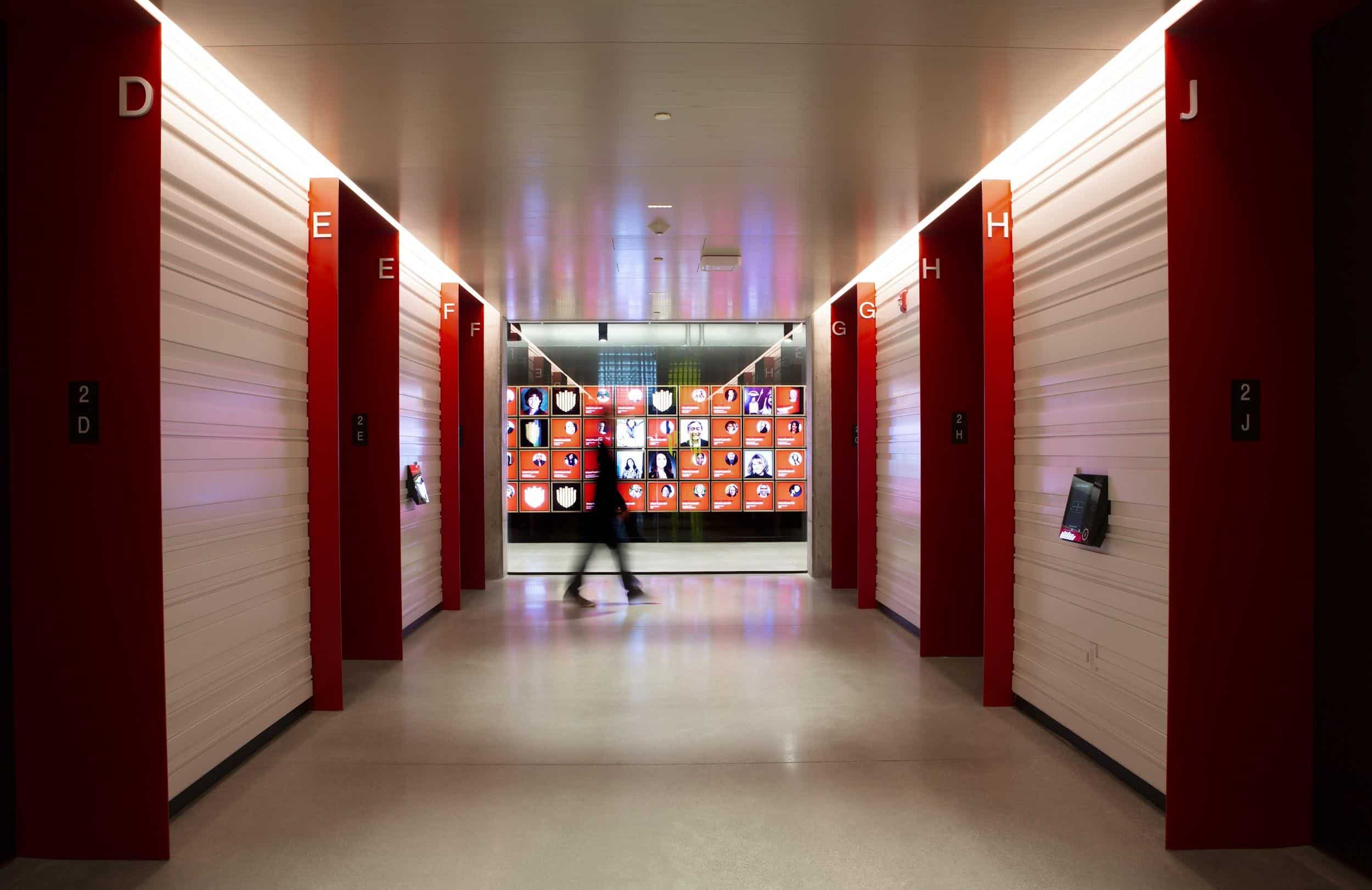 A person walks down a bright corridor with red walls, polished floor; screens ahead. Corridor labeled D to J.