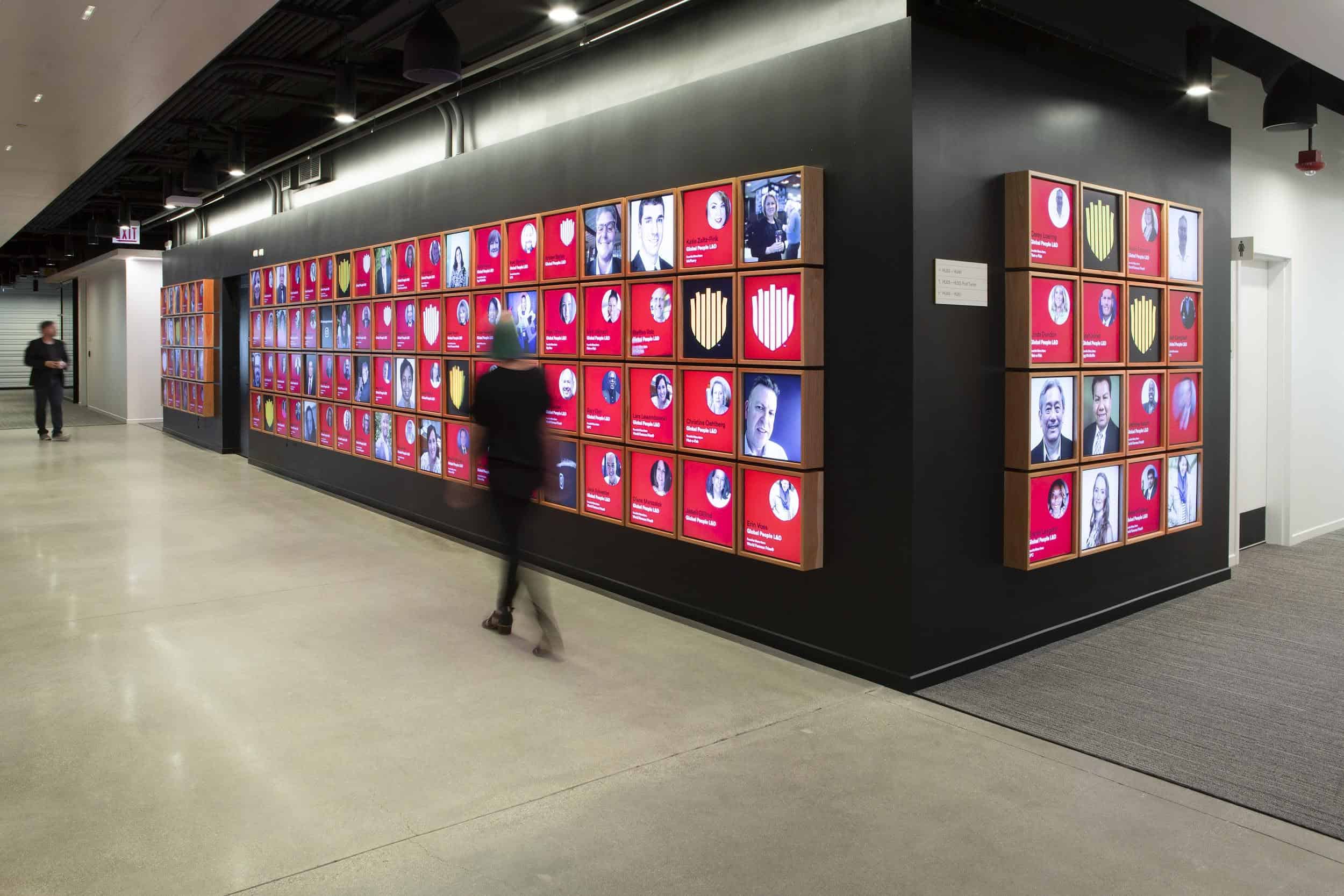 A person walks by a wall of framed photos with red and blue accents, in a corridor with polished concrete and exposed ductwork.