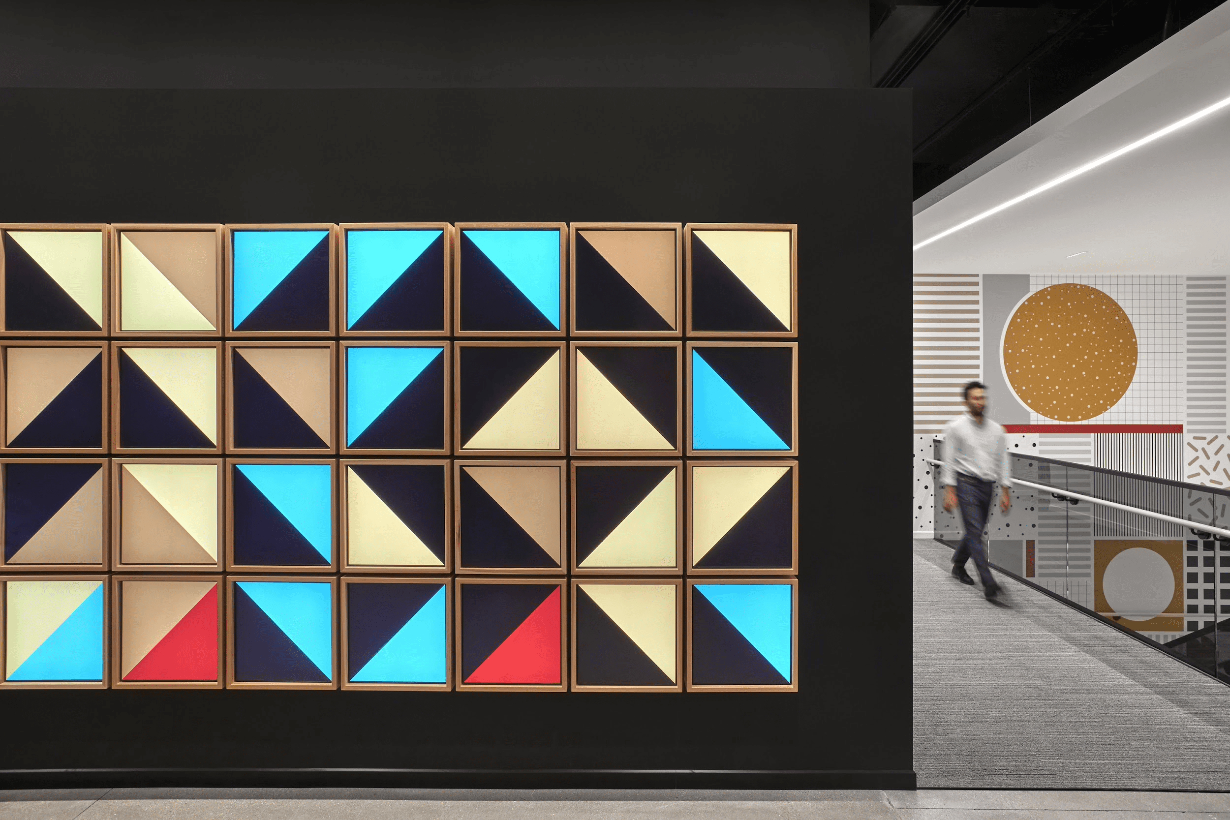 Colorful geometric wall art: squares split into black, blue, gold, cream triangles. Person walks past; abstract art in background.