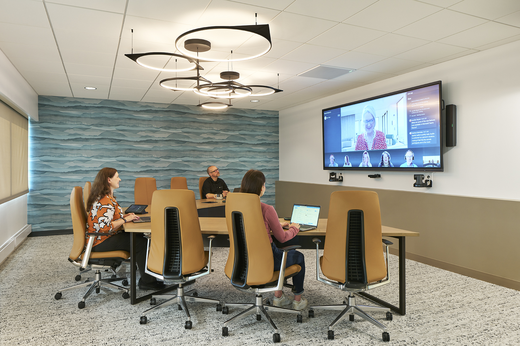 Three people in a modern conference room on a video call; wave-patterned wall, circular ceiling lights.