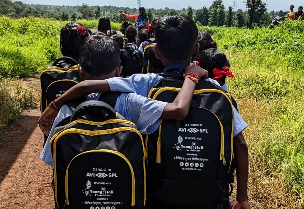 Kids in uniforms walk closely through greenery, carrying black backpacks. Two in front embrace, showing camaraderie.