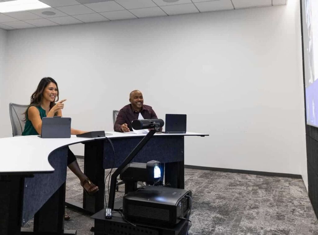 Two people at a curved desk with laptops; one smiles, the other gestures to a large screen. A projector is in front of them.