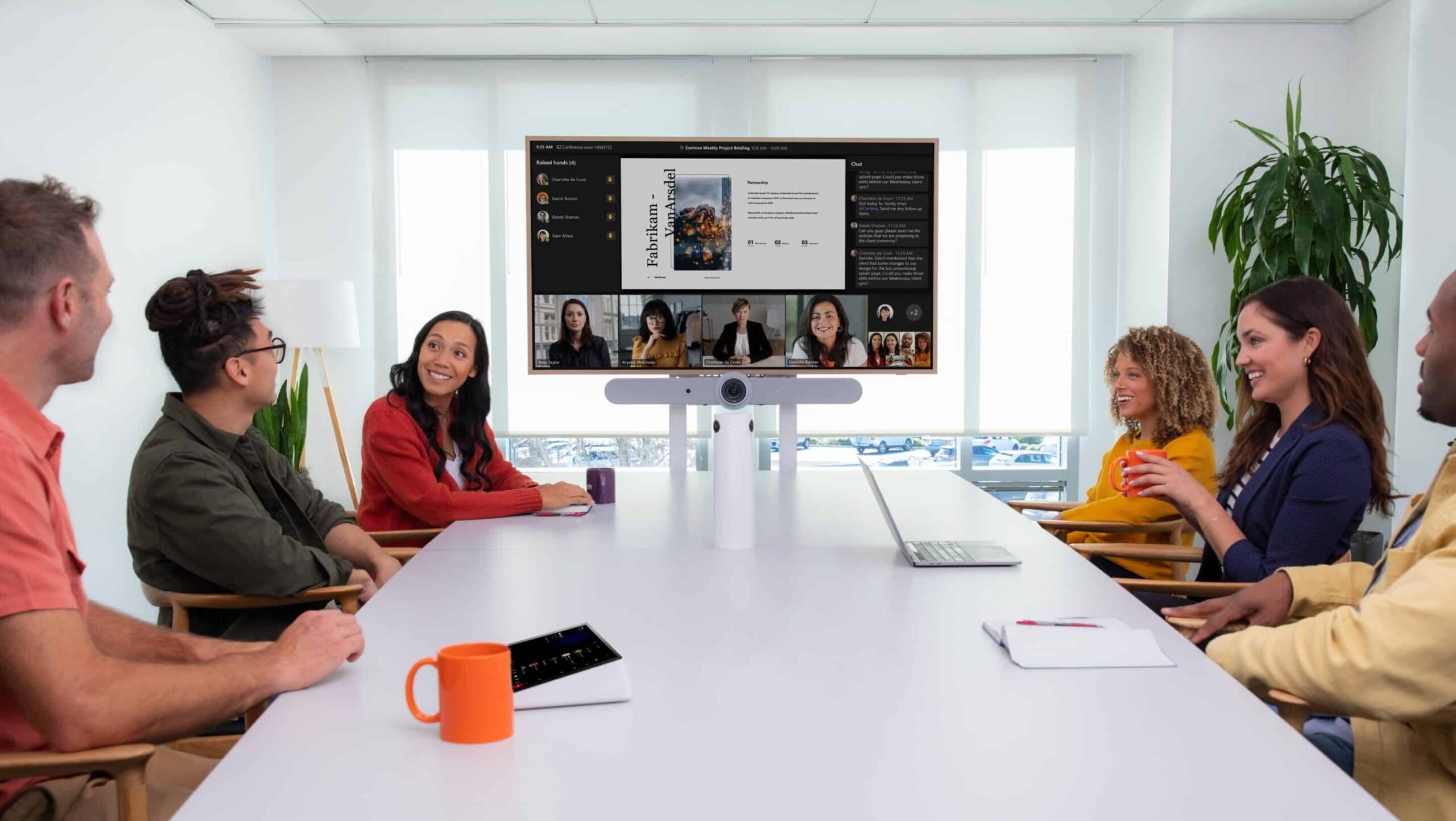 Six people sit around a table, engaging in a video call. Laptops, mugs, a potted plant, and natural light fill the room.