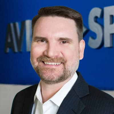 Smiling man in a dark suit and white shirt, standing before a blue background with partial letters visible.