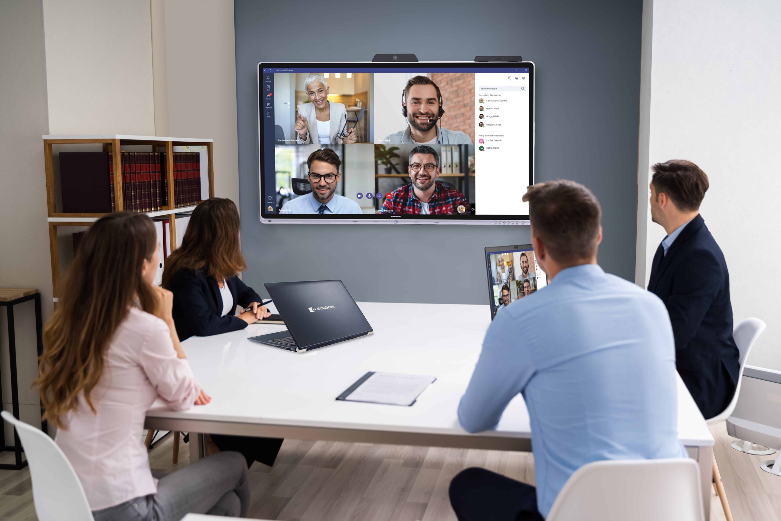 Four people in a conference room have a video meeting, laptops on the table; bookshelves visible in the background.