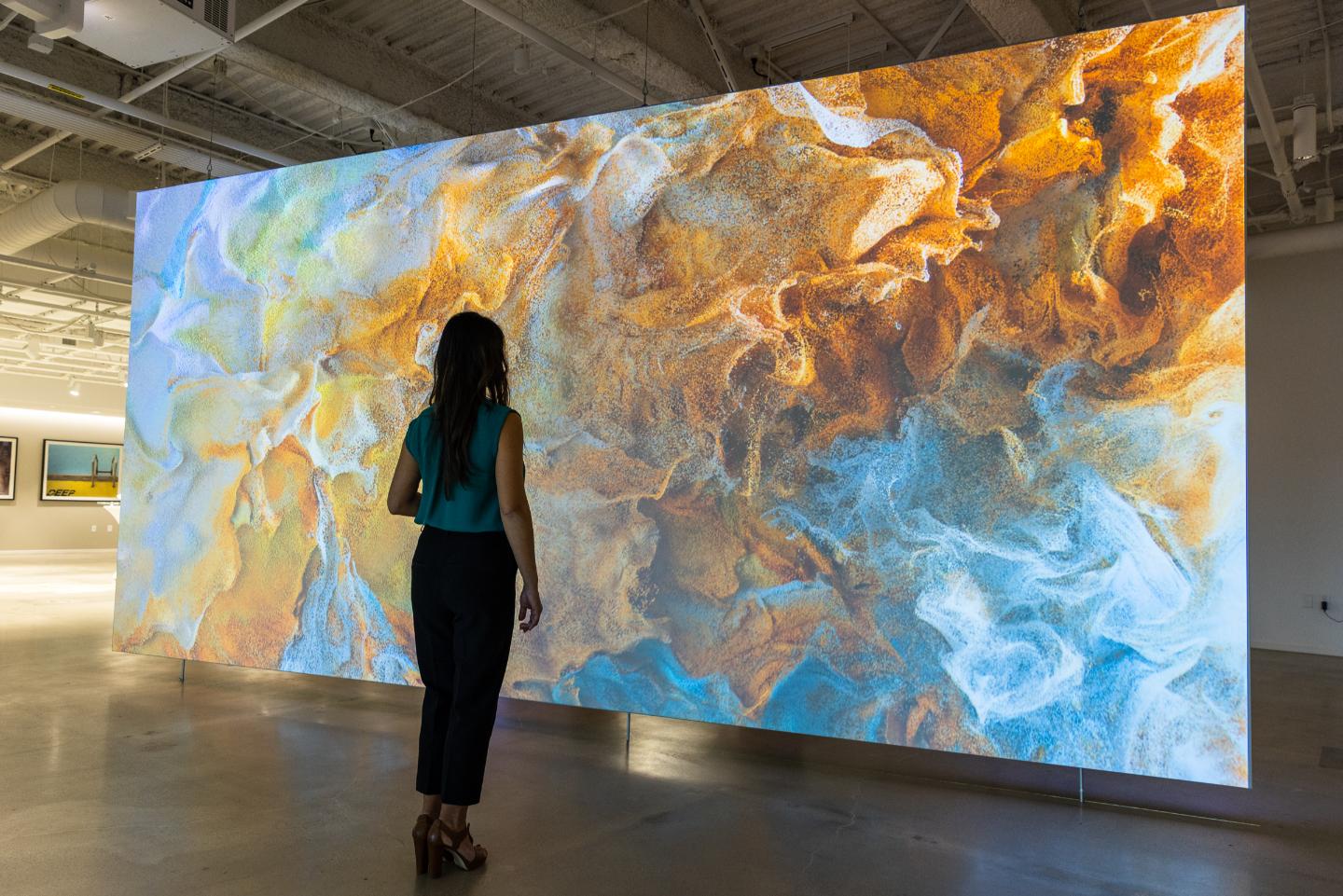 A person stands before a digital art display of swirling blue, orange, and white patterns in a spacious gallery.